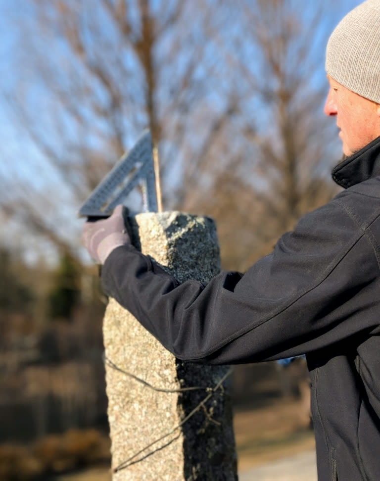 Close view of a gloved hand steadying a speed square on top of a weathered granite post